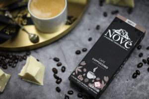 A box of Nove luxury chocolates resting next to a cappuccino on a grey slate background.
