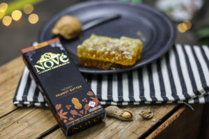 Budtender review Nove chocolates, ranking Honey Peanut Butter as one of their favorites.  The chocolate box rests on a wooden surface next to honeycomb and loose peanuts.