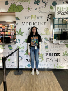 Abby, a budtender at Medicine Man dispensary, stands in front of their branded icon wall.